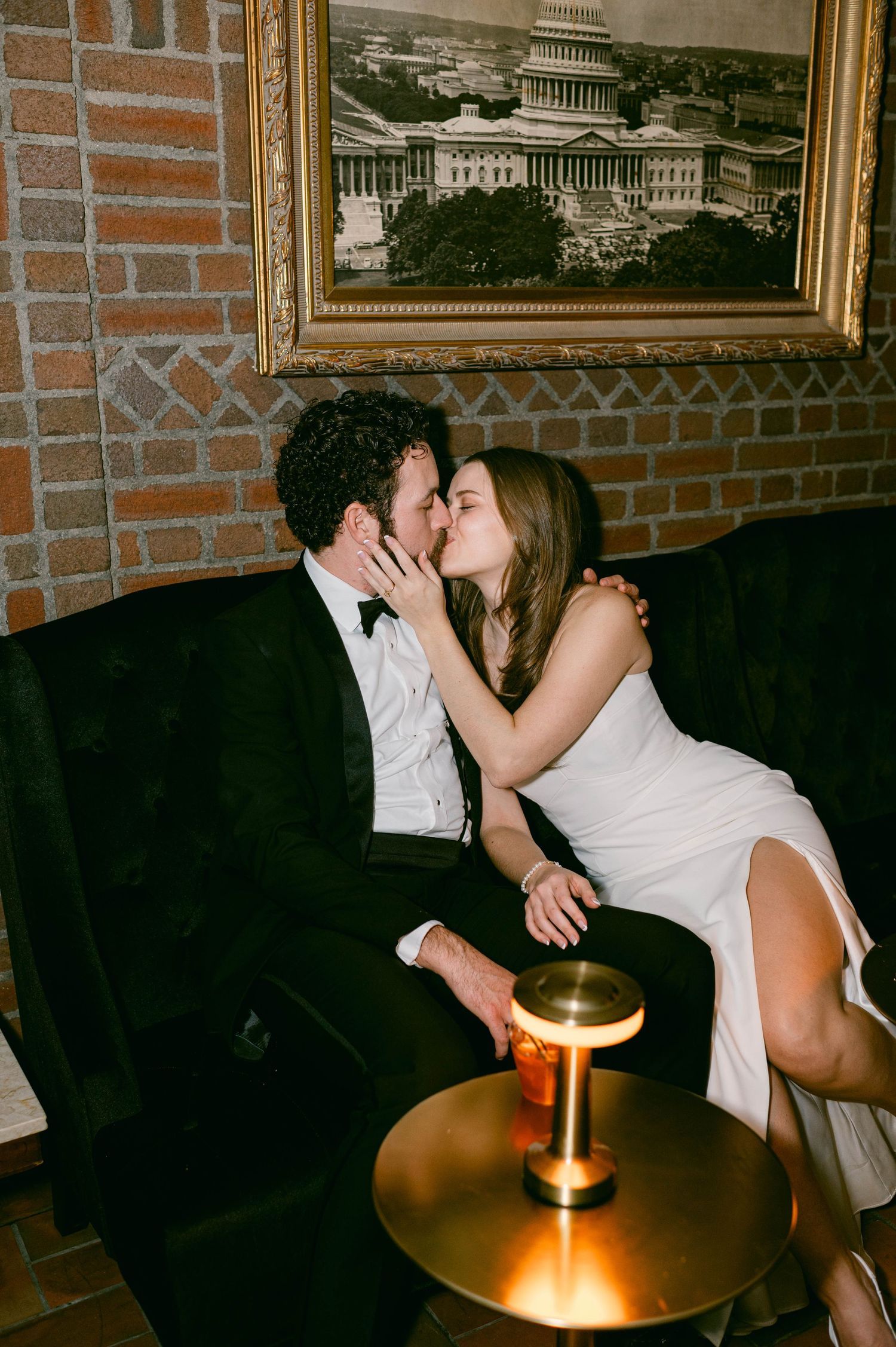 Couple in wedding attire sharing a romantic kiss inside a dimly lit room with exposed brick walls and framed artwork.