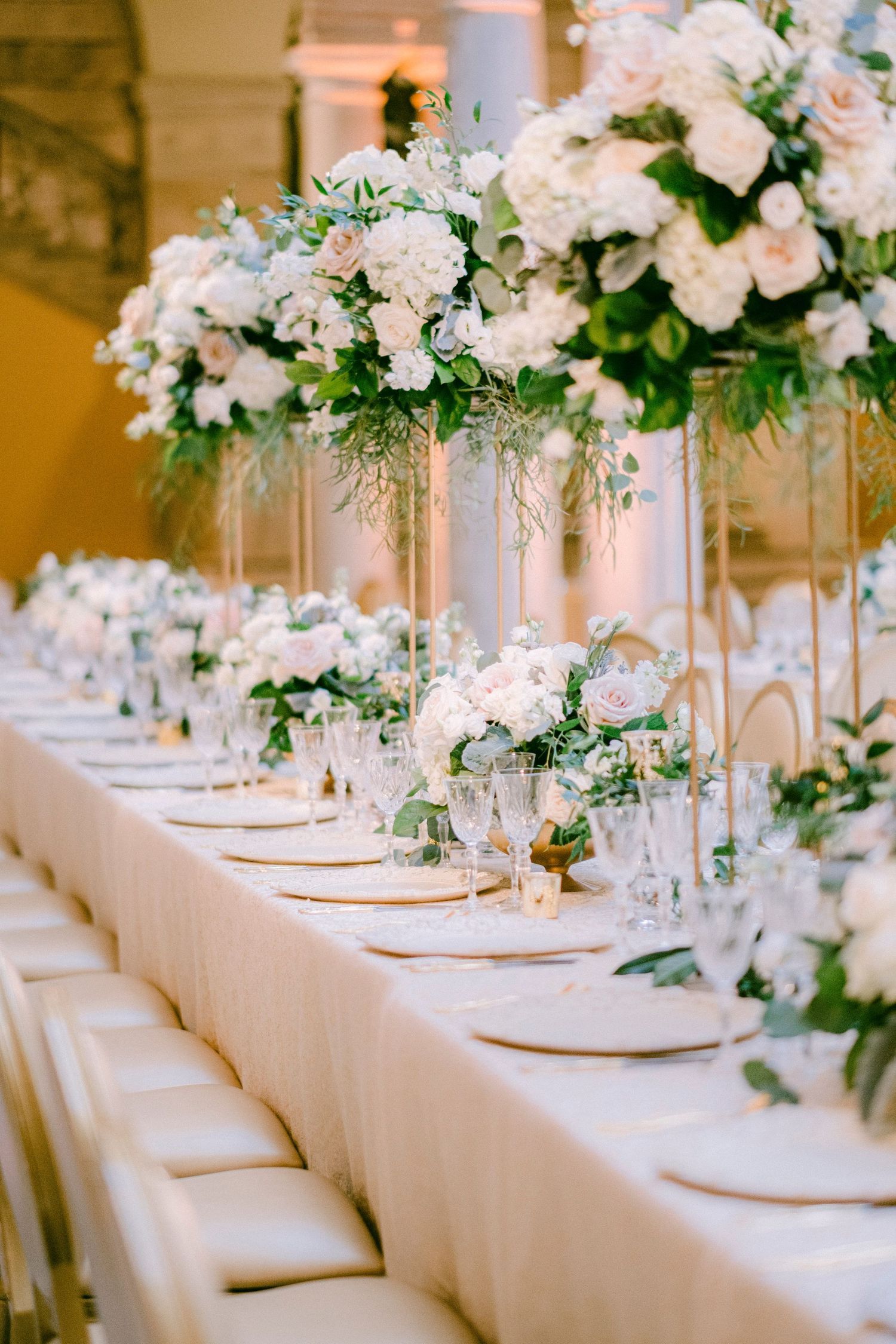 Elegant white floral centerpieces adorn a long wedding reception table with pristine place settings and white tablecloths.