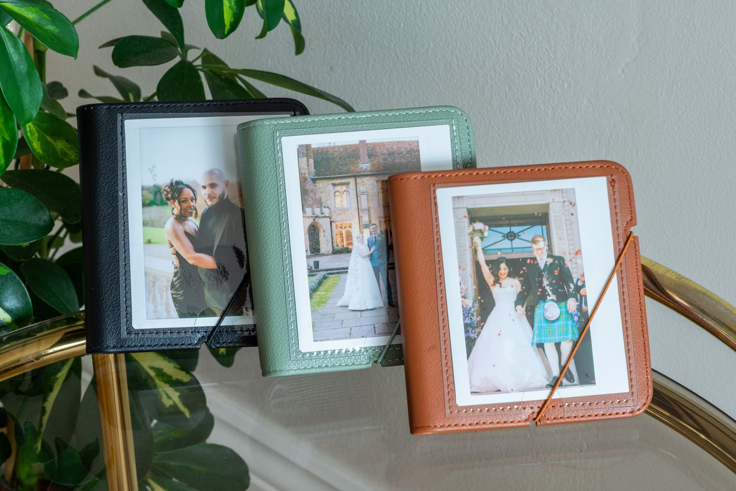 Wedding photo displays in mint green and brown leather frames sit on a brass table with greenery.