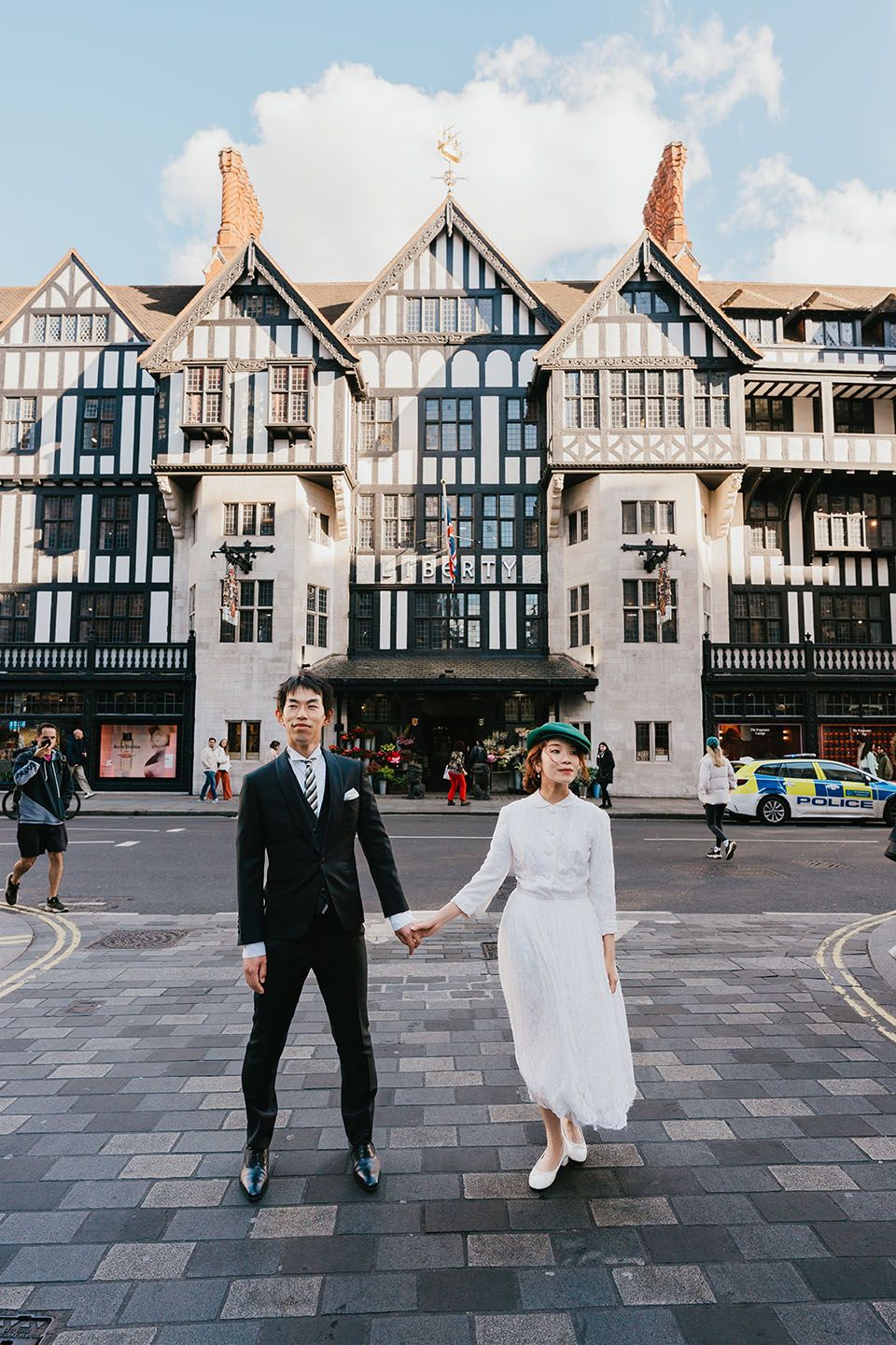 A couple poses in wedding attire in front of a Tudor-style building with timber framing in a British city street.