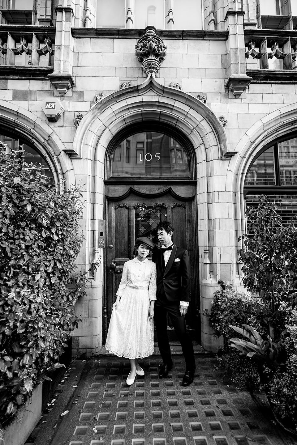 Vintage-style black and white photo of a couple in wedding attire standing in an ornate stone archway entrance.