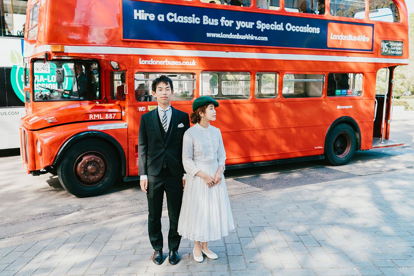 A couple poses in elegant attire in front of a vintage red double-decker London bus on a sunny day.