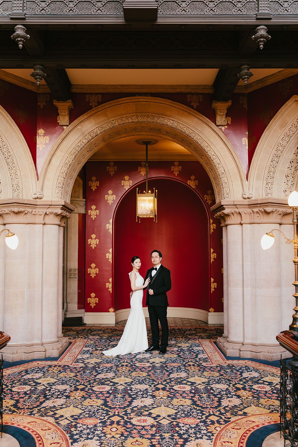 A couple stands in an elegant archway with ornate architectural details, red walls and a patterned carpet below.