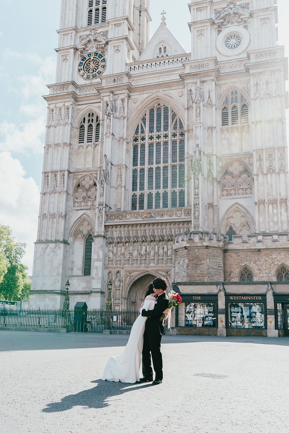 A couple embraces in front of Westminster Abbey's grand Gothic architecture on a bright sunny day in London.