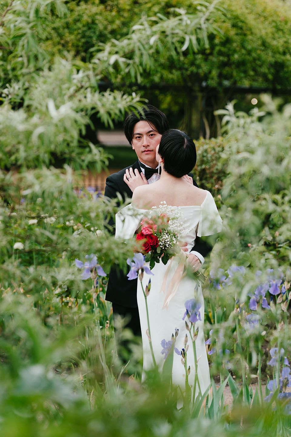 A couple embraces in a romantic garden setting surrounded by purple irises and lush greenery.