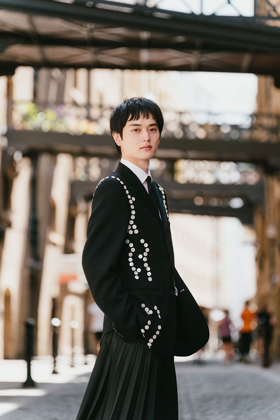 A person in a black outfit with decorative pearls poses under an urban bridge structure in soft lighting.