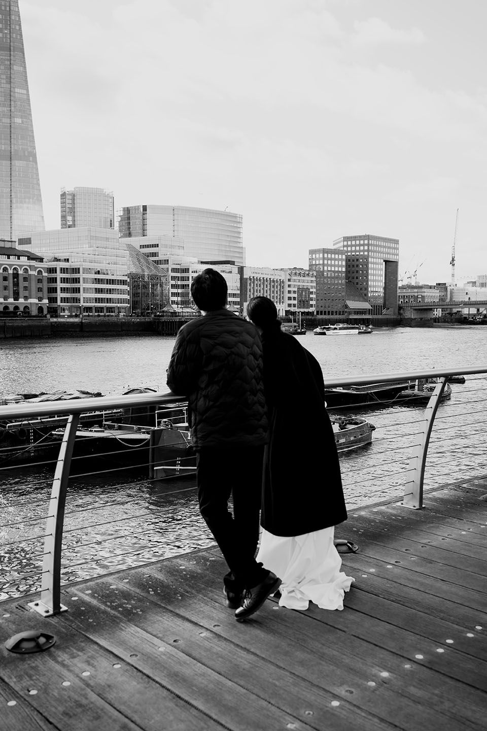 Two people stand together on a wooden pier overlooking the River Thames with London's modern skyline in the background.