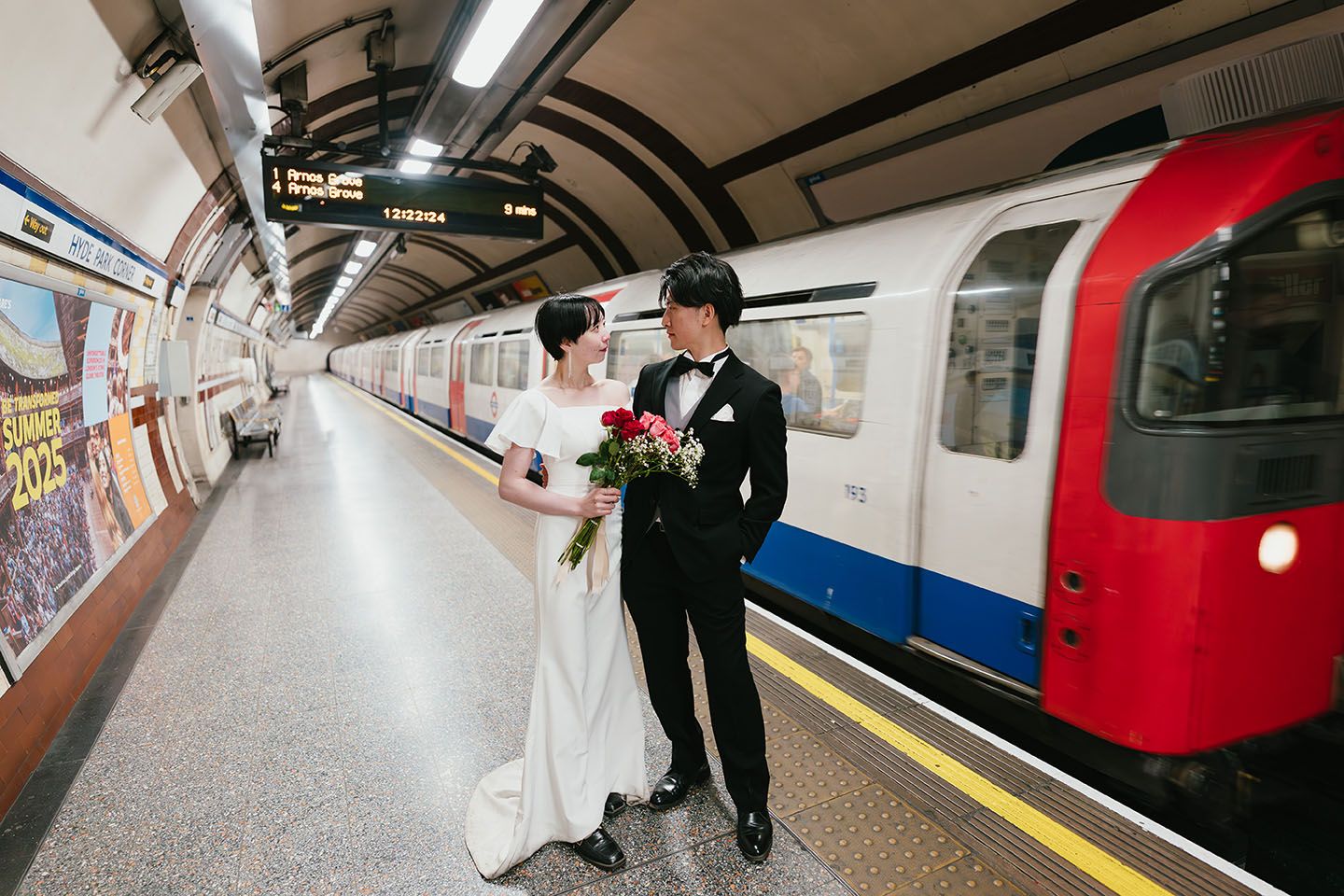 A bride and groom stand together on an empty London Underground platform with a red and blue train visible.