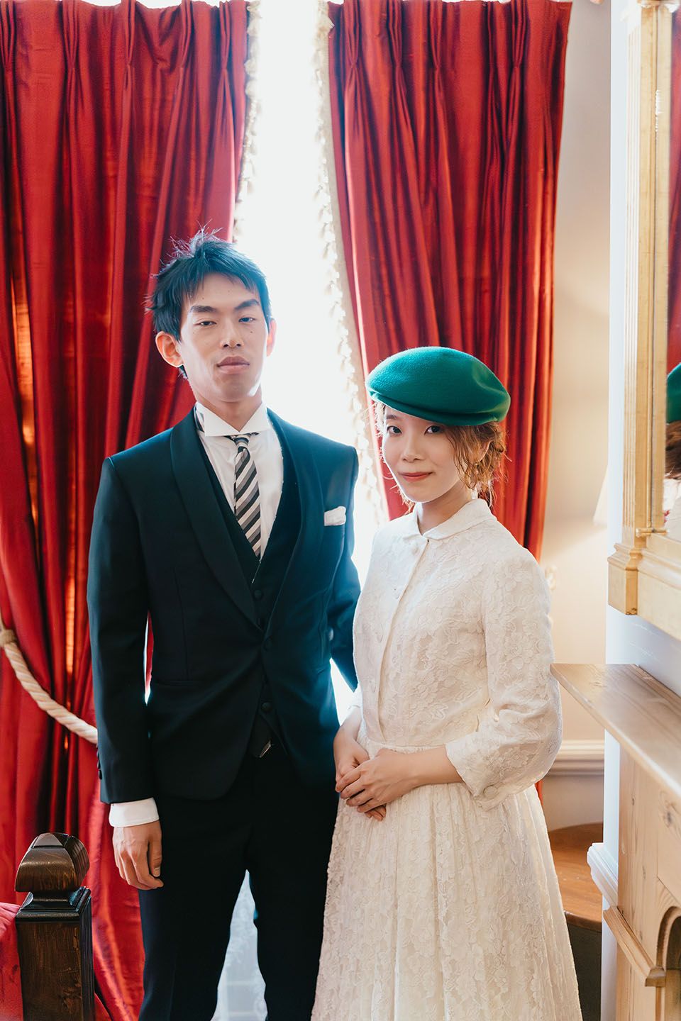 A couple in vintage-inspired formal wear pose together against red curtains, with a distinctive green beret accent.