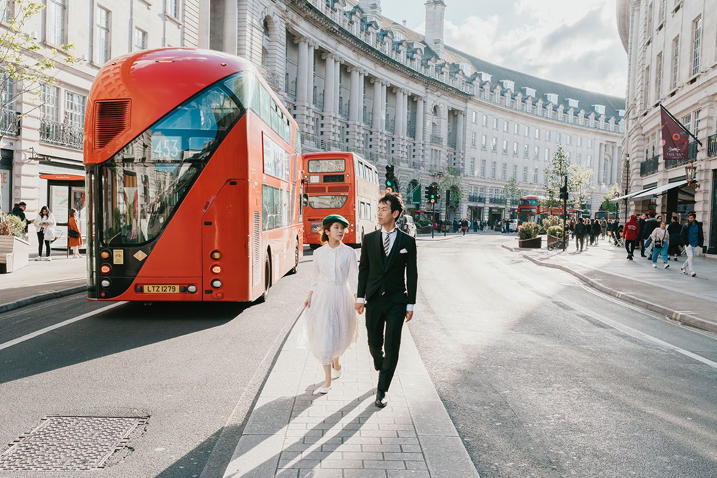 A bride and groom walking past a red London double-decker bus on a city street.