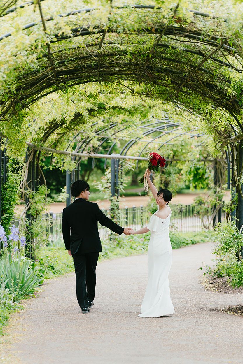 A couple walks hand in hand under a lush green garden archway adorned with climbing vines and white flowers.