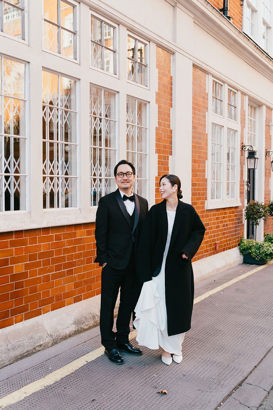 A couple in formal attire stands together against a brick building with white windows in an elegant urban setting.
