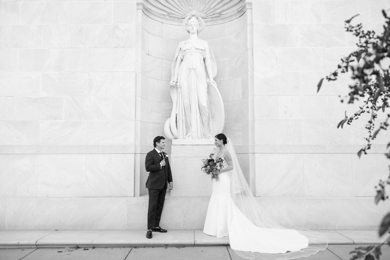 A bride and groom pose in front of a large white marble statue in an elegant architectural setting.