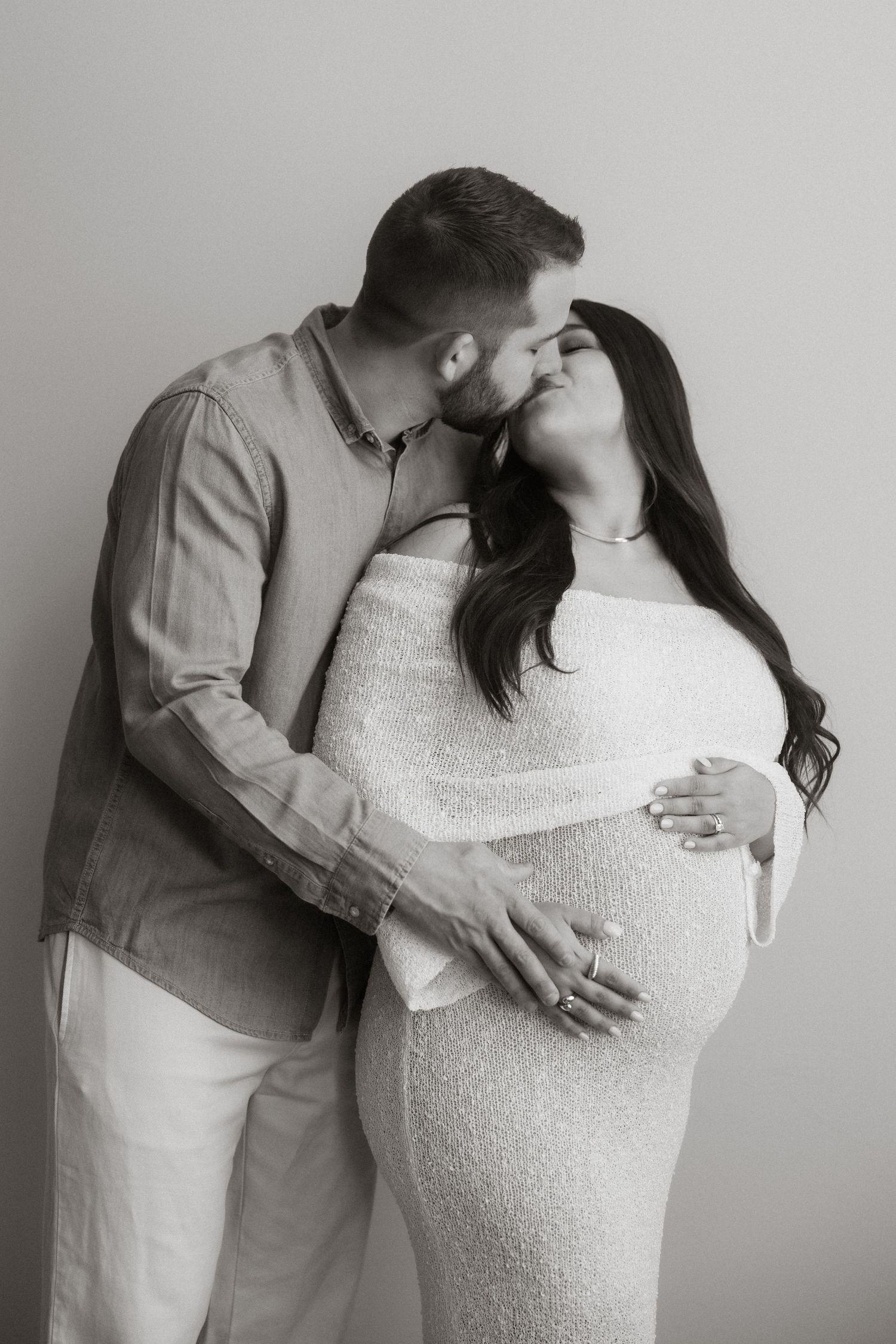 Black and white maternity portrait showing intimate moment between expectant couple in coordinated light-colored outfits.