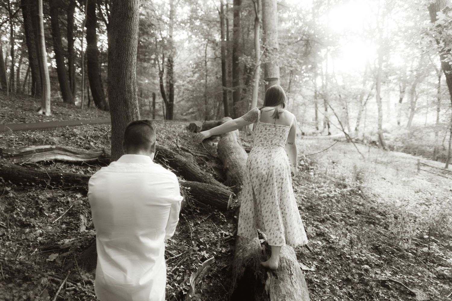 A black and white artistic wedding photo shows two people in formal attire on a wooded hillside trail.