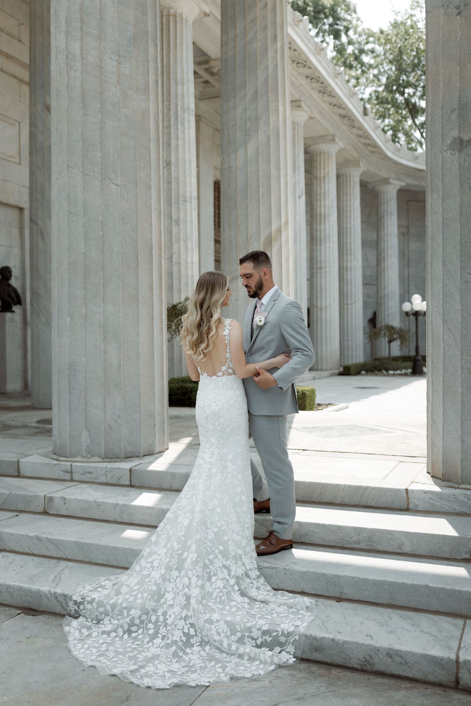 Wedding couple stands between elegant white columns, bride's lace train flowing down stone steps.