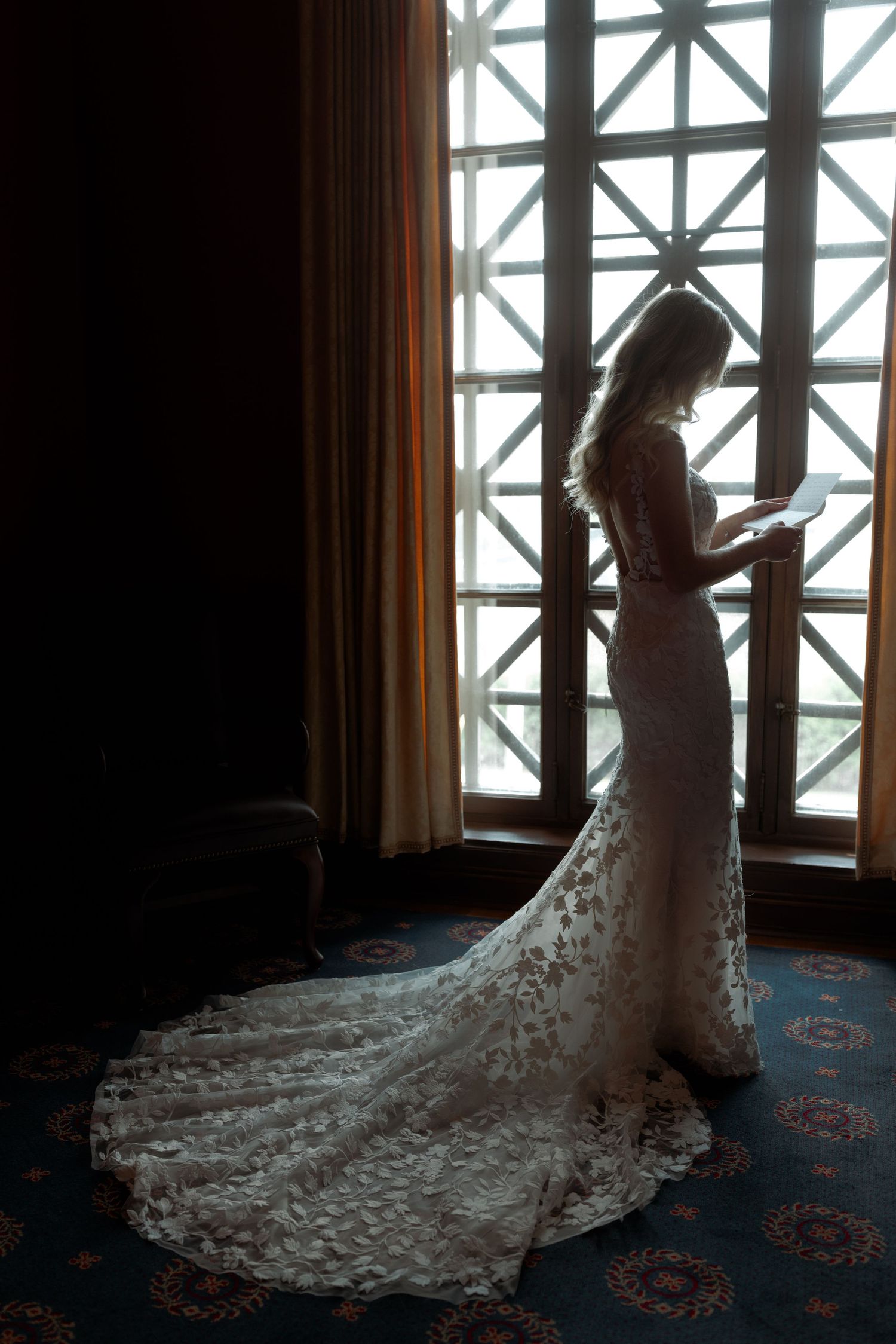 A silhouetted figure in a lace wedding dress with long train stands by a geometric window.