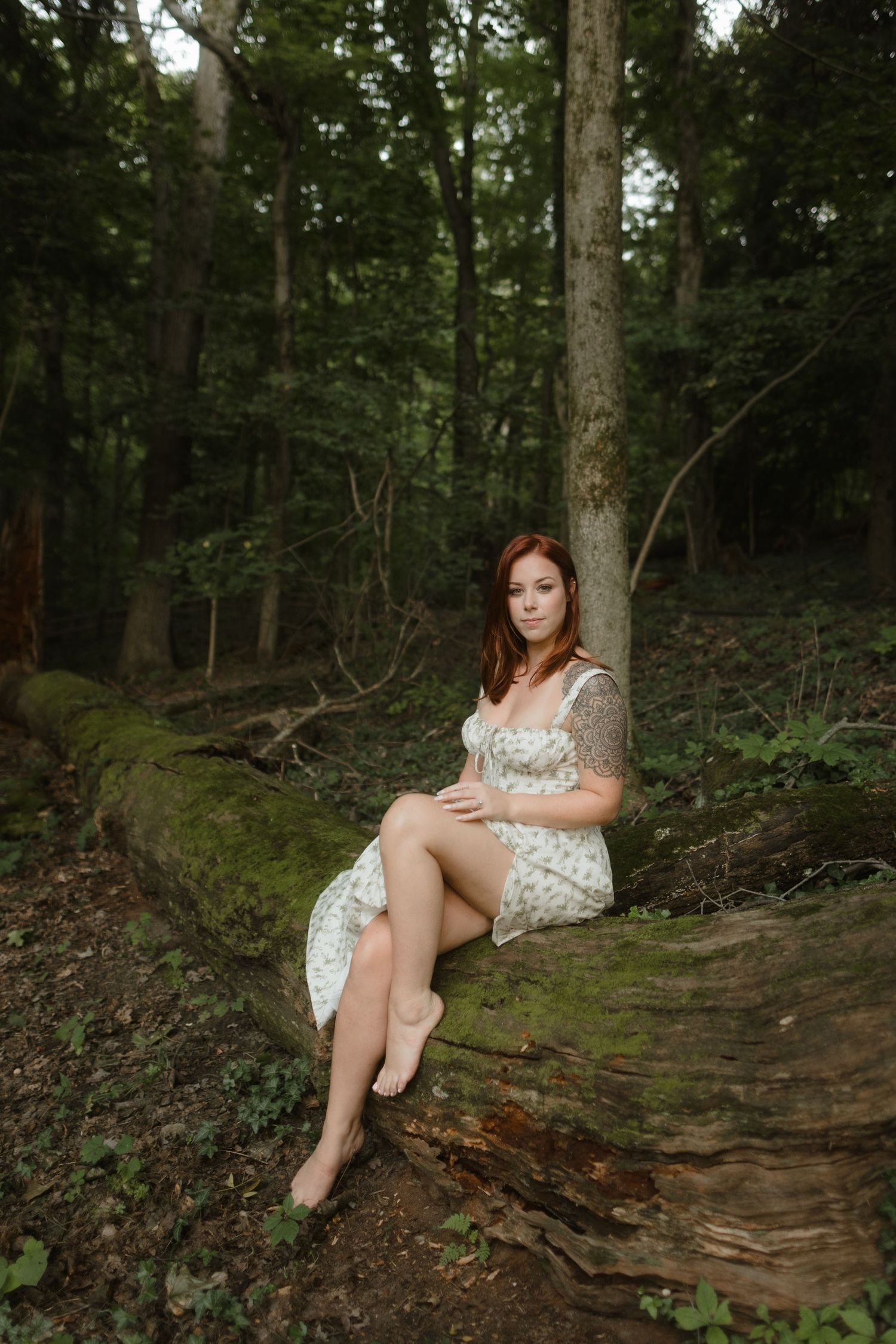 A figure in a white floral dress lounges thoughtfully on tree-covered forest terrain during a photo session.