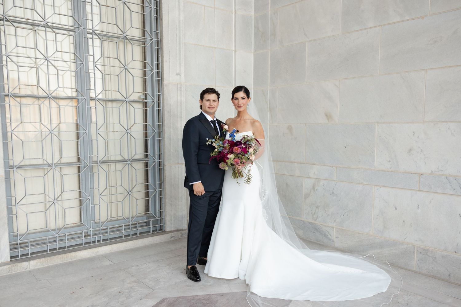 A bride and groom walking together under the white stone arches of an elegant architectural corridor.