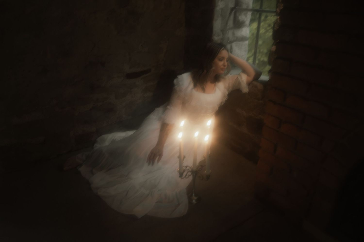 Ethereal figure in flowing white dress sits on wooden floor illuminated by soft candlelight in dark moody setting.