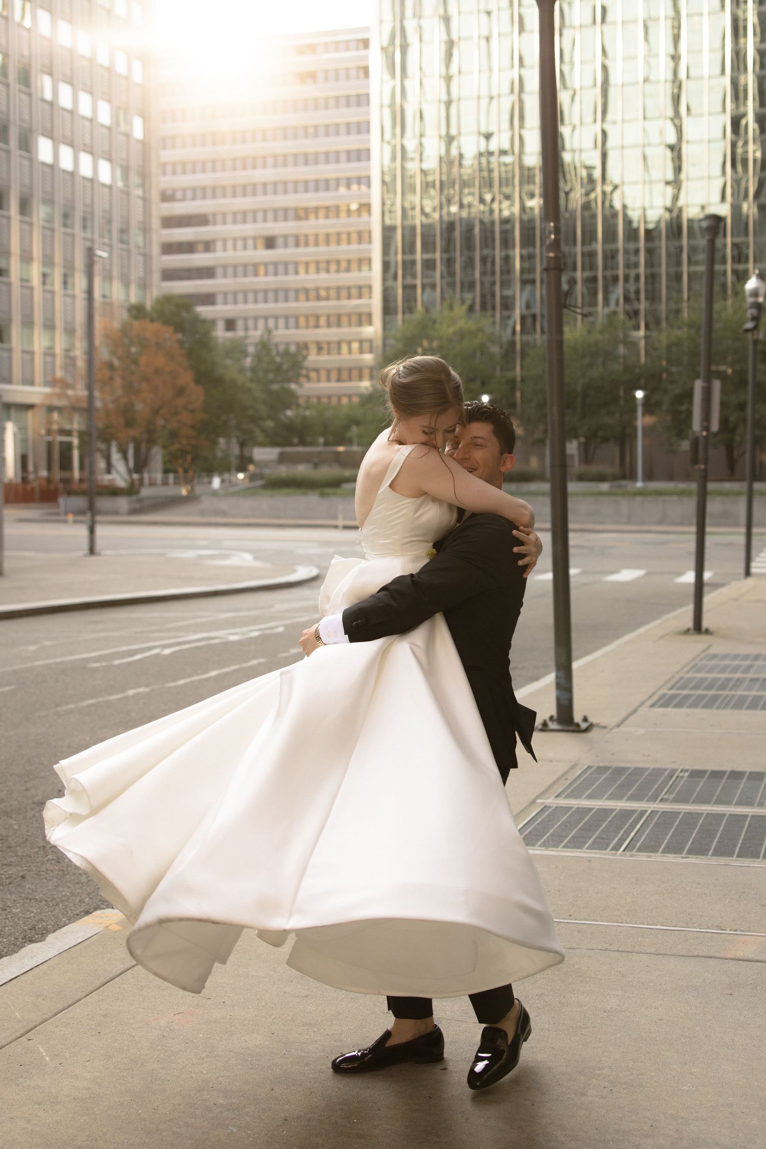 Groom lifts bride in flowing dress during sunset dance on city street.