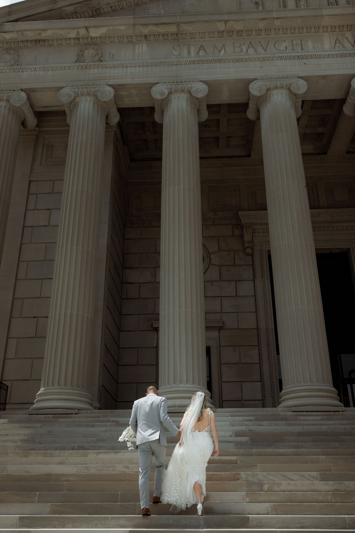 A couple ascending stone steps of a grand building with classic Greek-style columns in the background.
