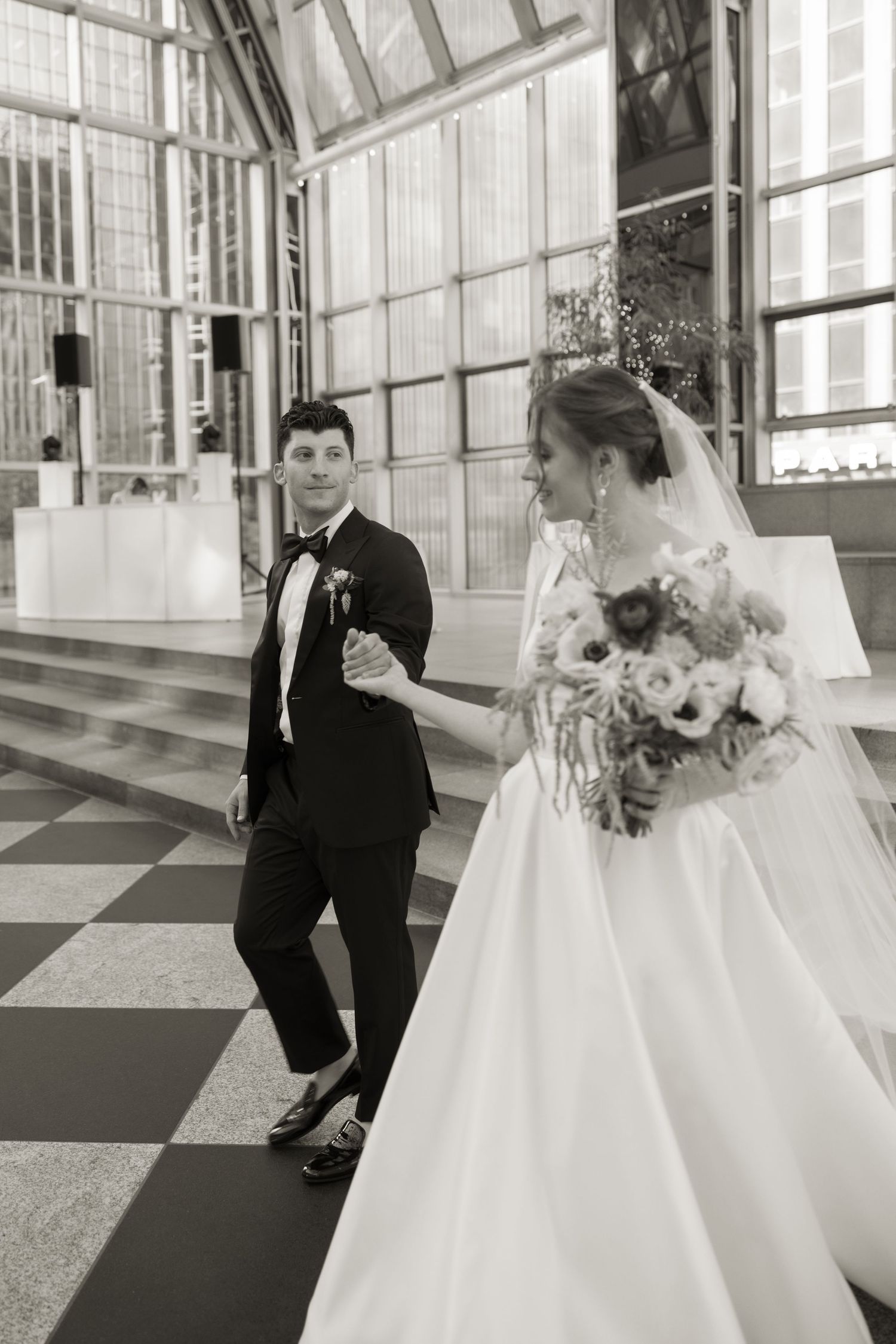 Black and white photo of newlyweds walking together on checkered floor in glass atrium.