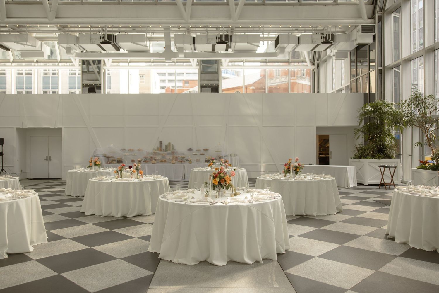 Modern wedding reception space with white round tables on checkered floor and natural lighting.