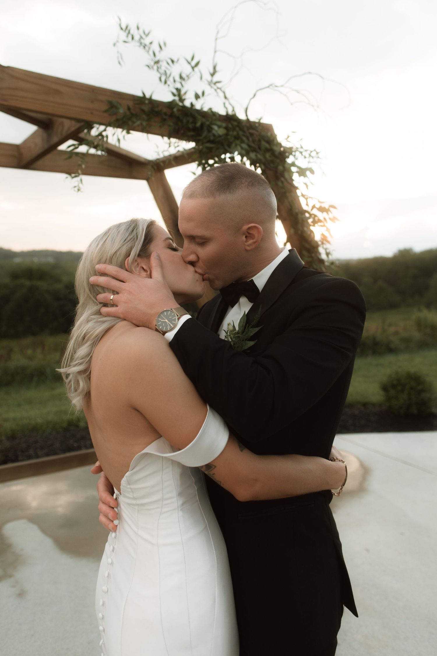 A romantic wedding kiss under an arch decorated with greenery at sunset.