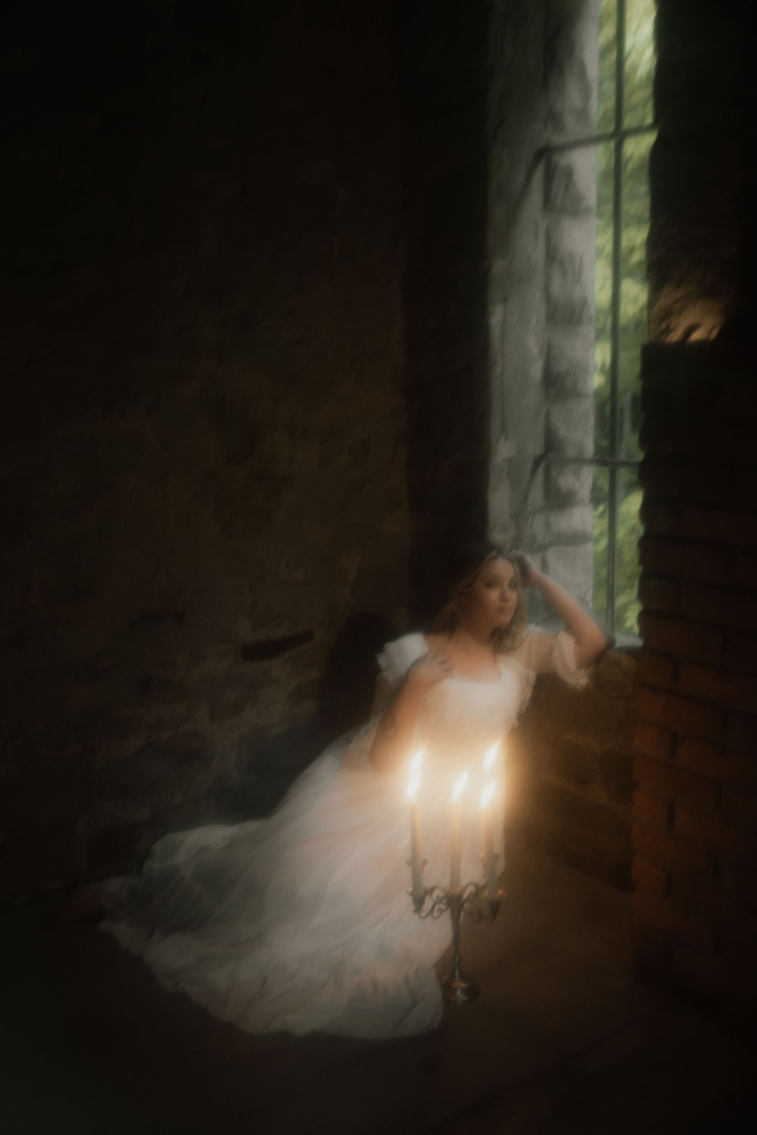 Ethereal figure in flowing white dress sits near window in dark moody room creating dreamy romantic atmosphere.