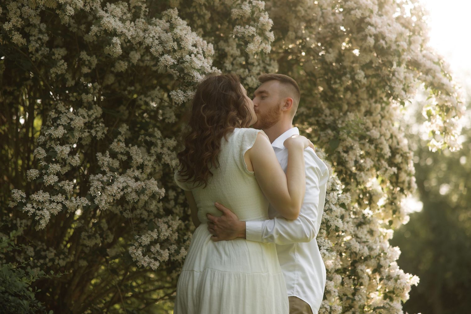 A couple sharing a romantic kiss in front of blooming white flowers during golden hour sunlight.