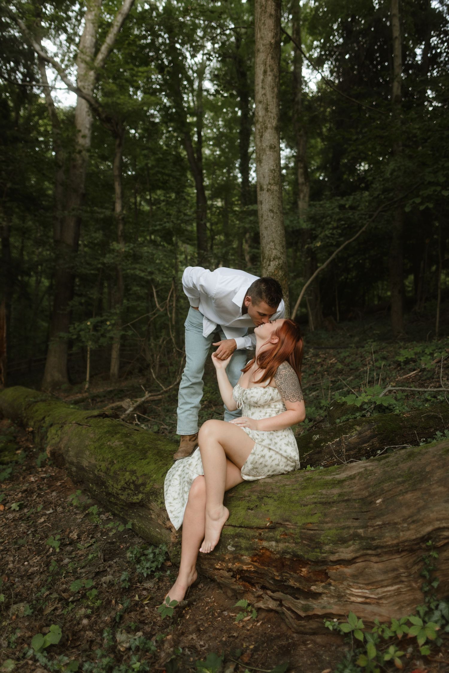 A playful couple shares a tender moment on a fallen log in a wooded forest setting.