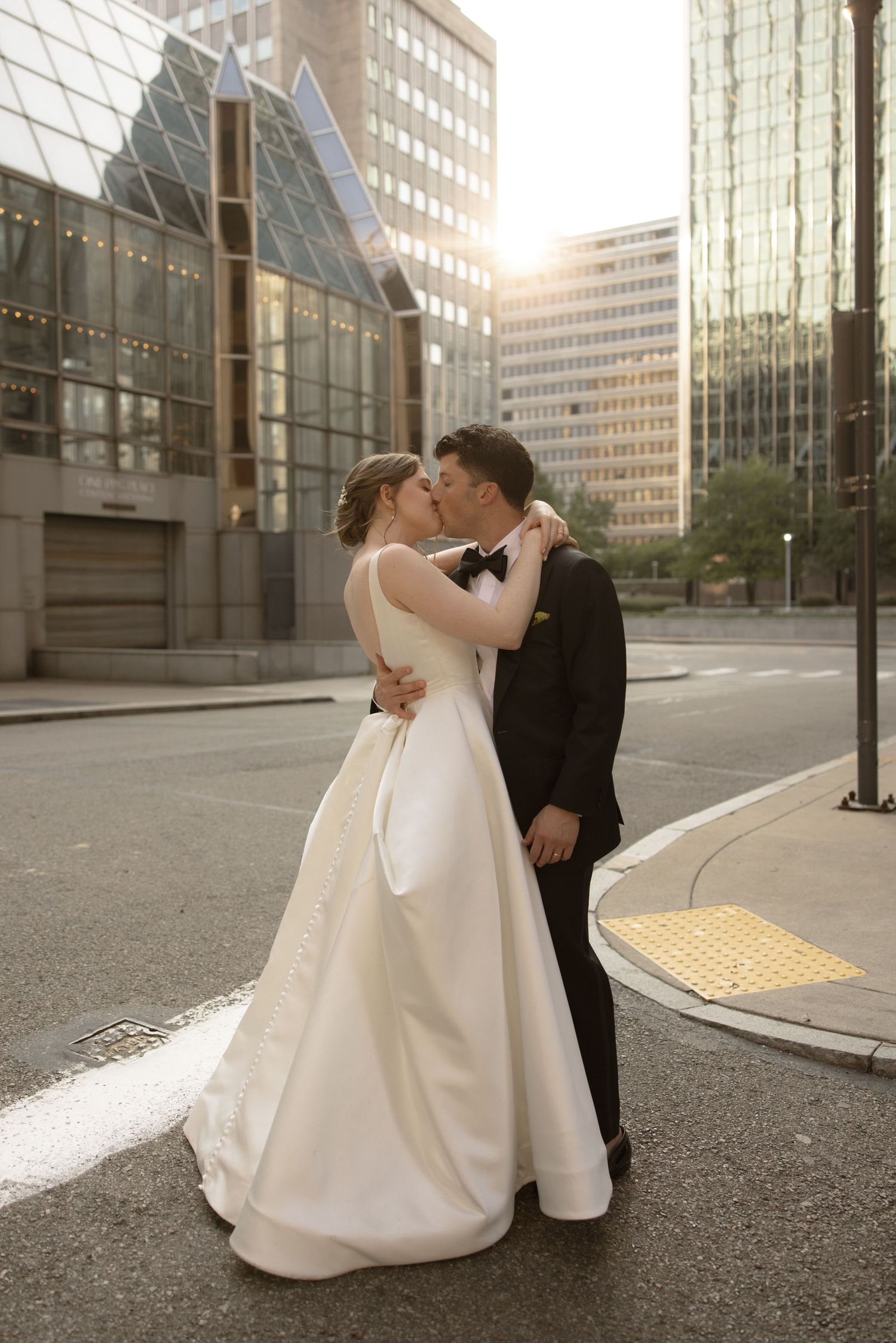 Wedding couple shares a romantic kiss on city street with modern architecture in background at sunset.