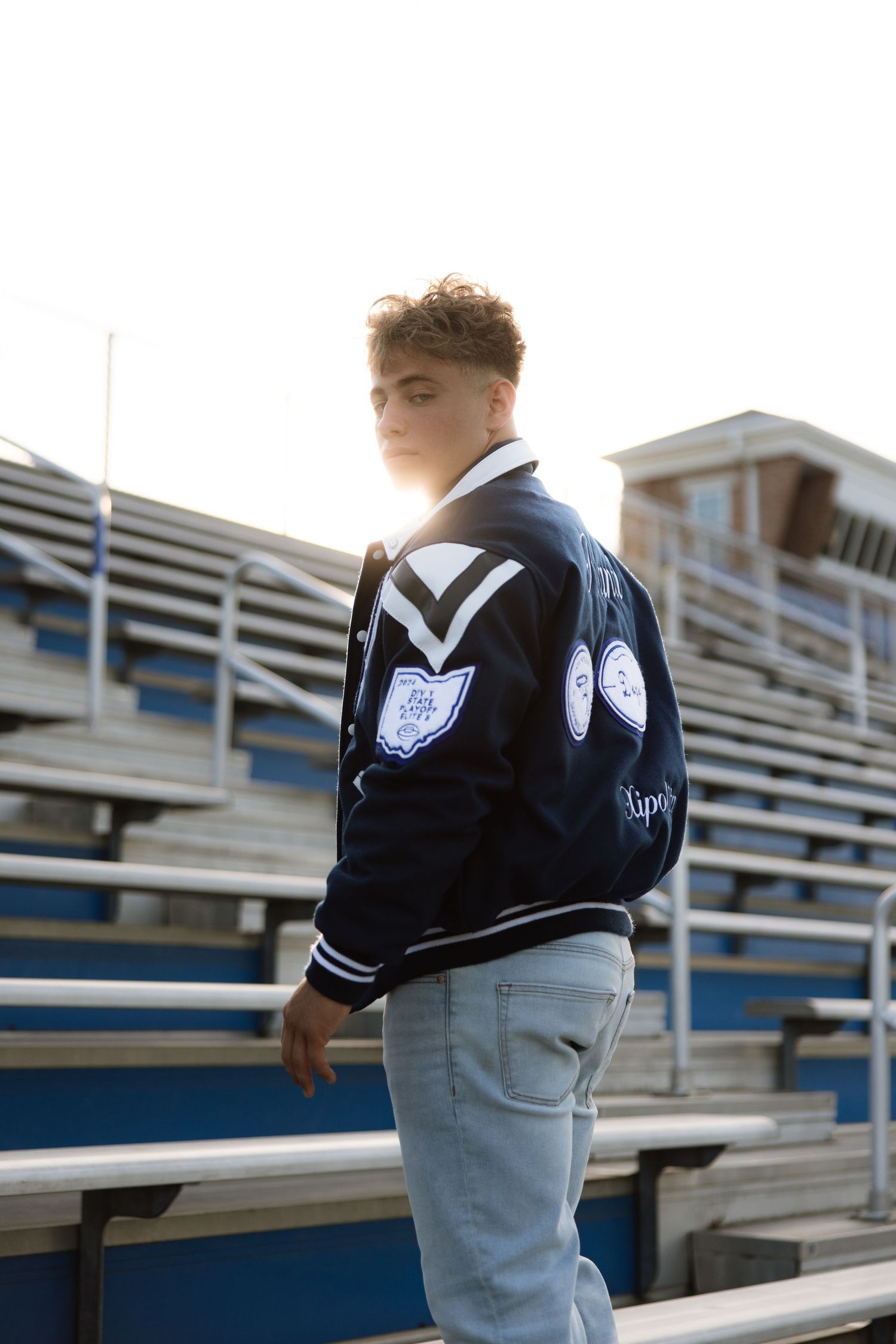 Student in varsity letter jacket poses on bleachers at sports stadium.