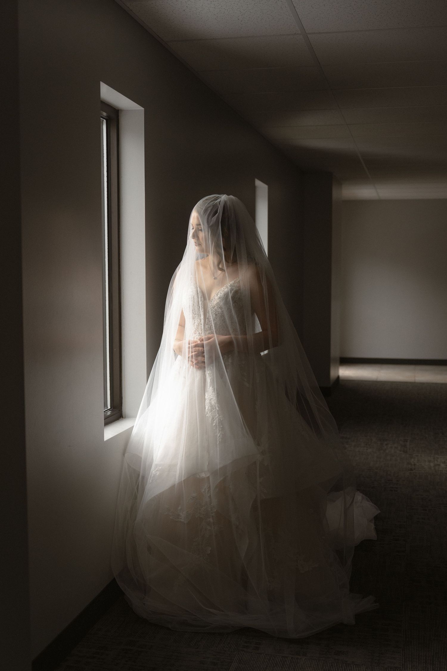 A bride in a flowing white wedding gown and long veil stands silhouetted against a bright window in a dark hallway.
