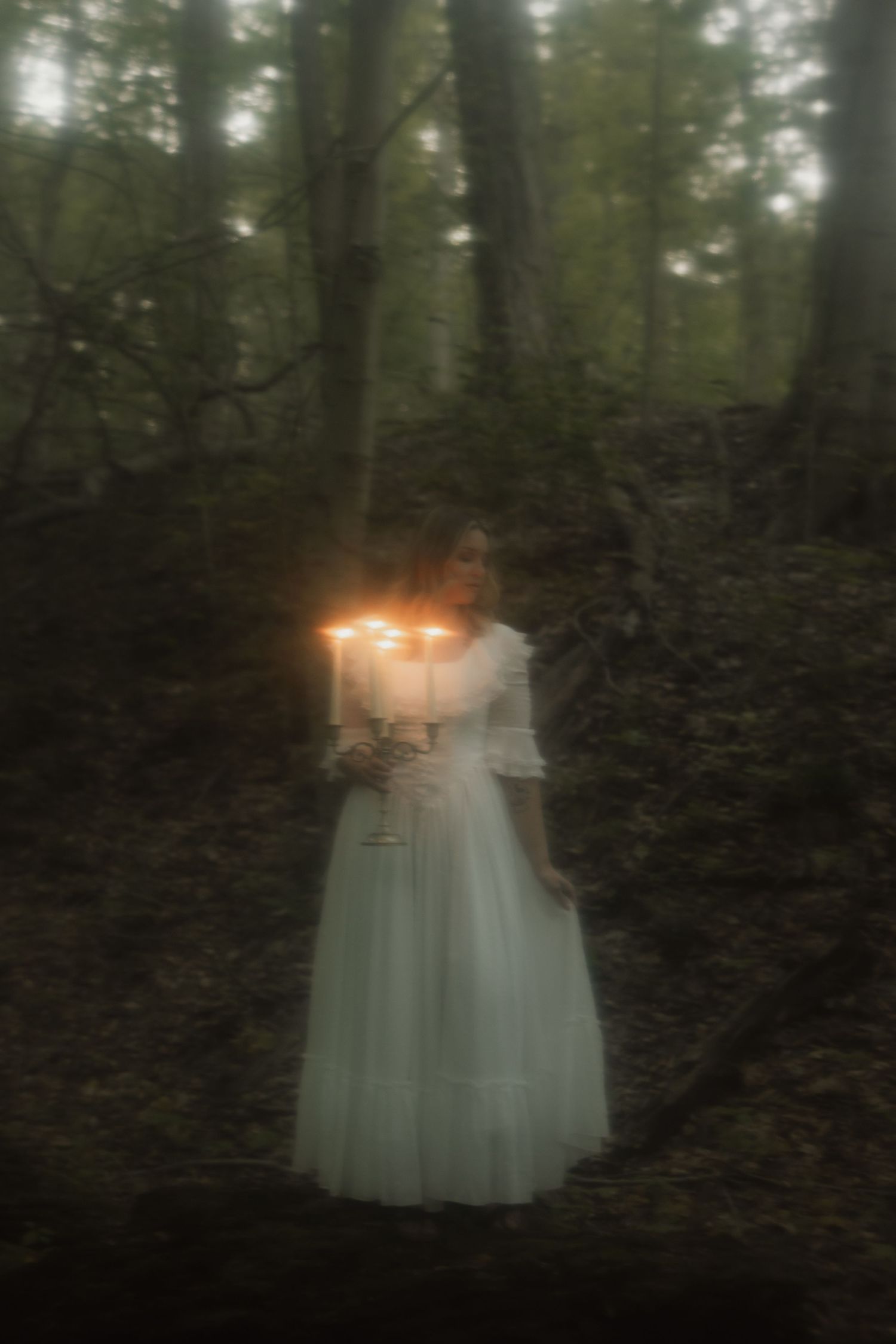 A mysterious figure in a white dress holds glowing candles in a misty, dark forest creating an ethereal atmosphere.