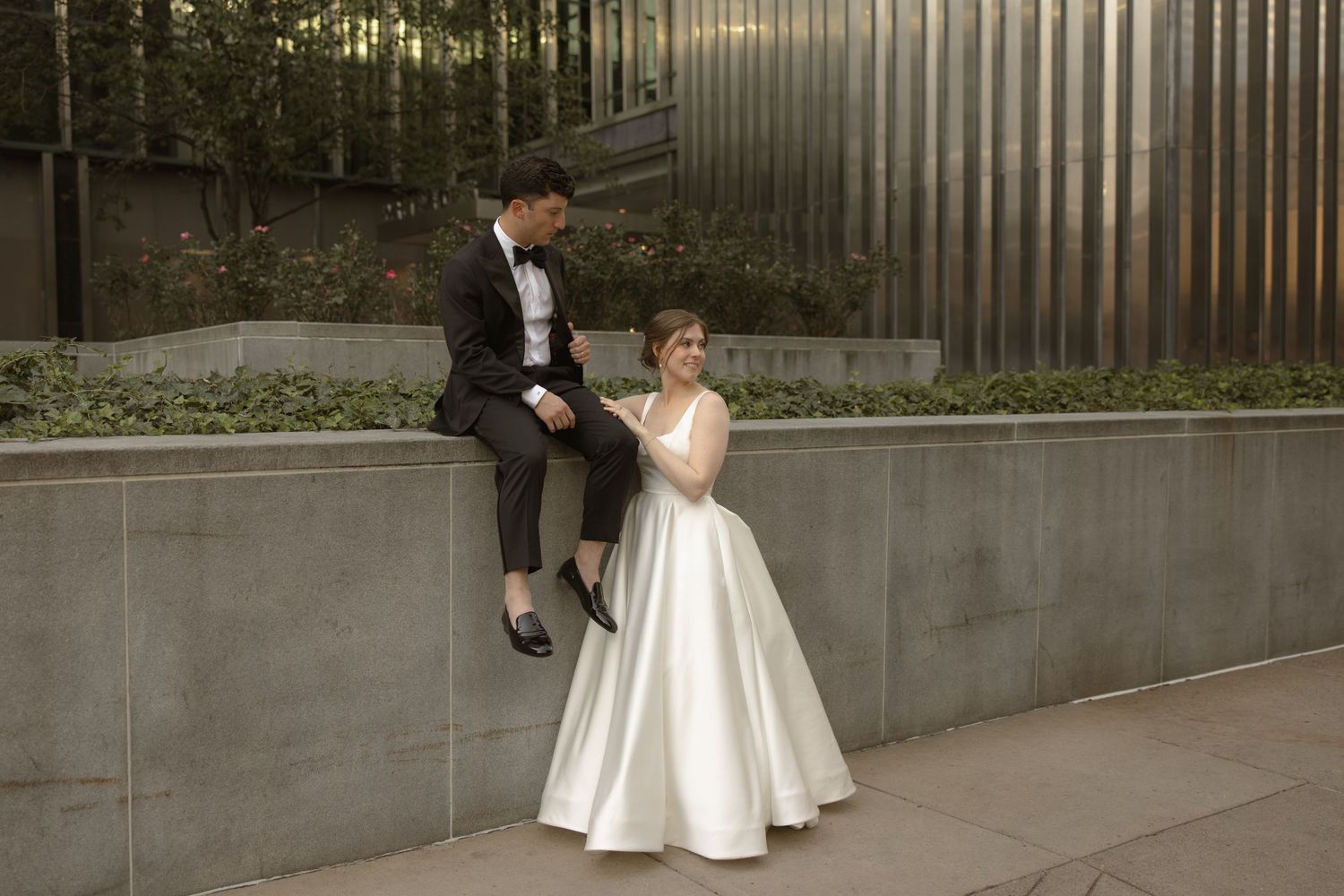 Newlyweds share a casual moment sitting on concrete wall in urban setting.