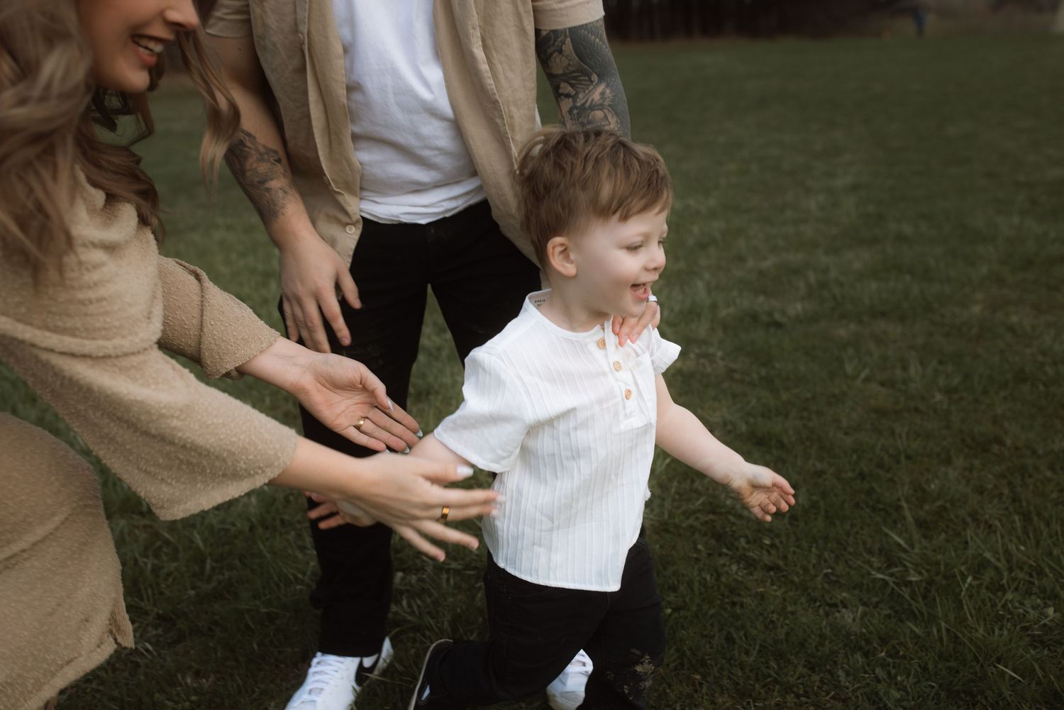 A toddler in white running and playing with parents on grass.