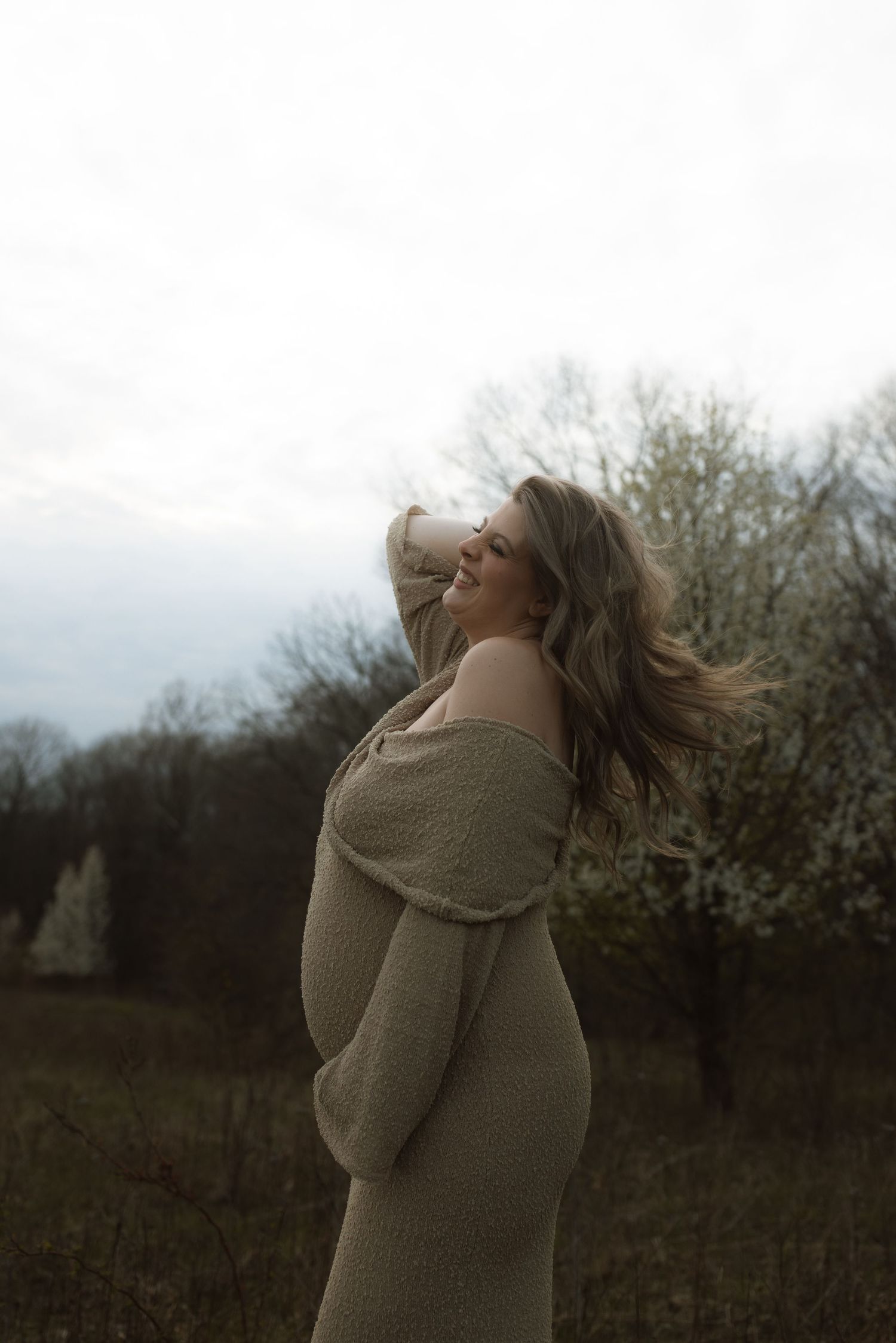 Expectant woman in beige sweater dress poses joyfully in a winter field.