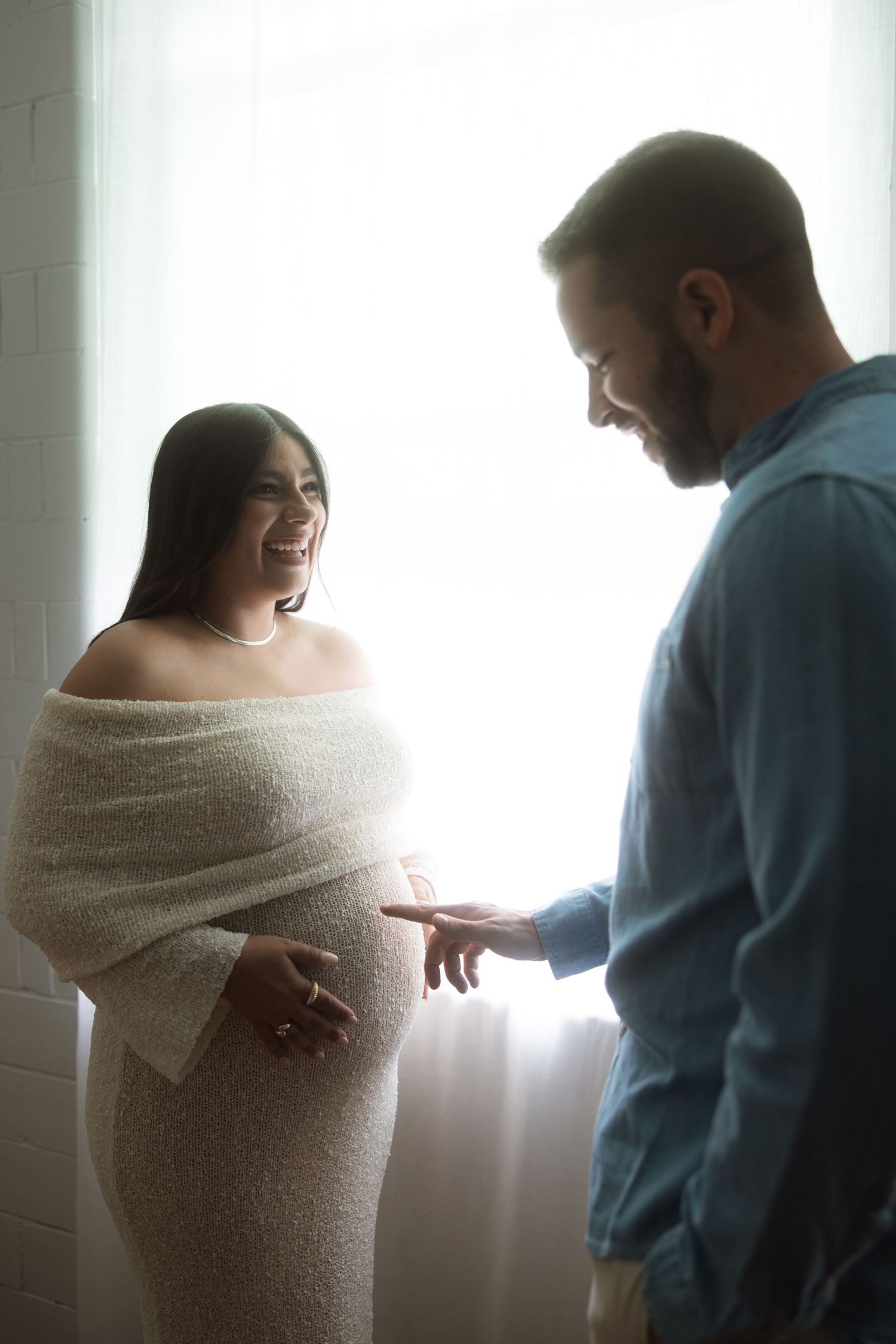 A romantic moment with a softly lit couple standing intimately together by a bright window.