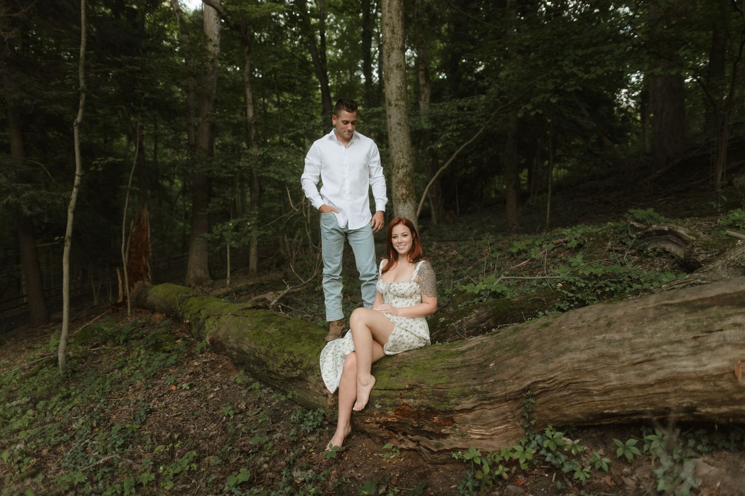 A couple in light colored clothing poses together on a fallen tree trunk in a wooded forest setting.
