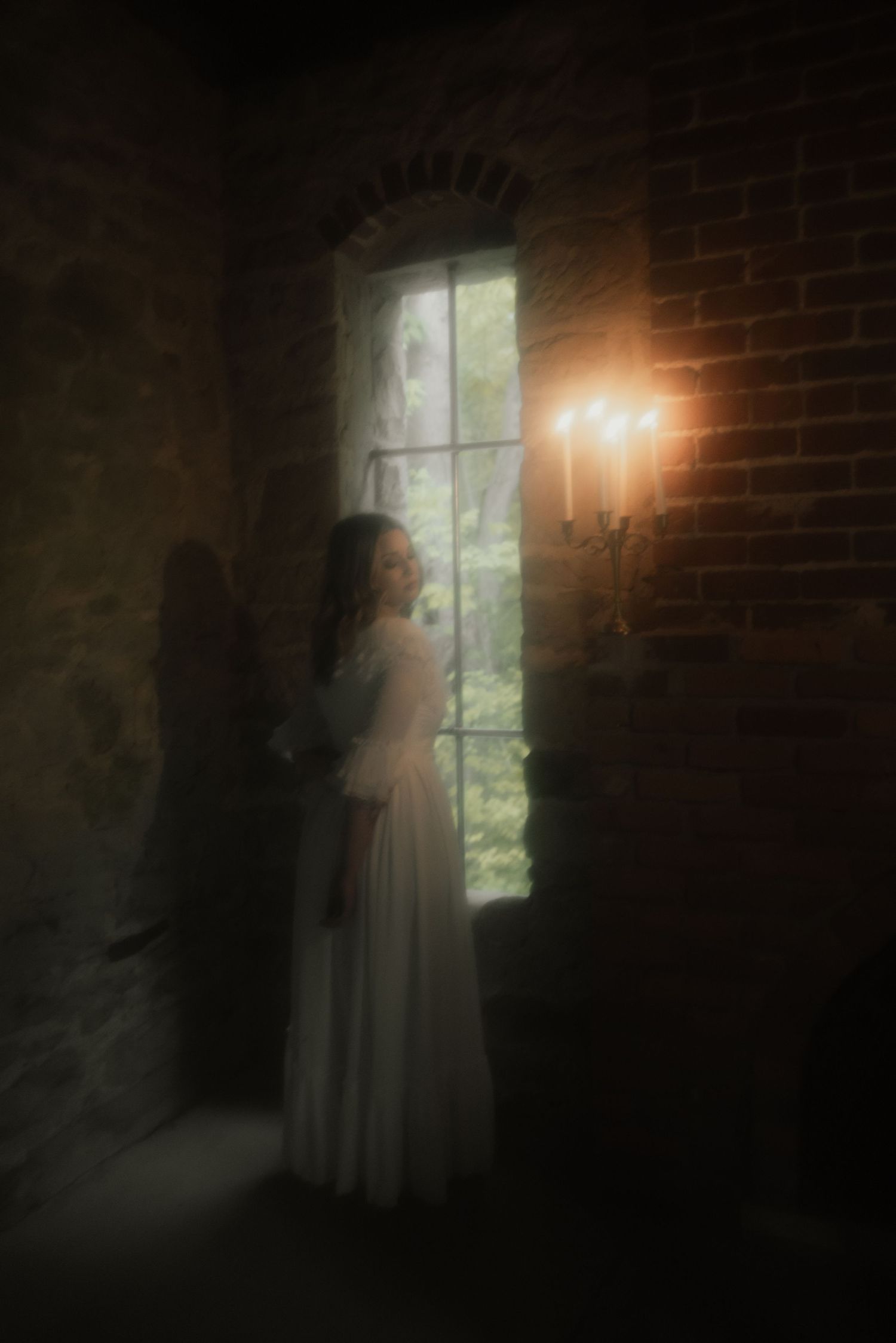 A figure in a flowing white dress stands near a candlelit window in a dimly lit brick room creating an ethereal atmosphere.