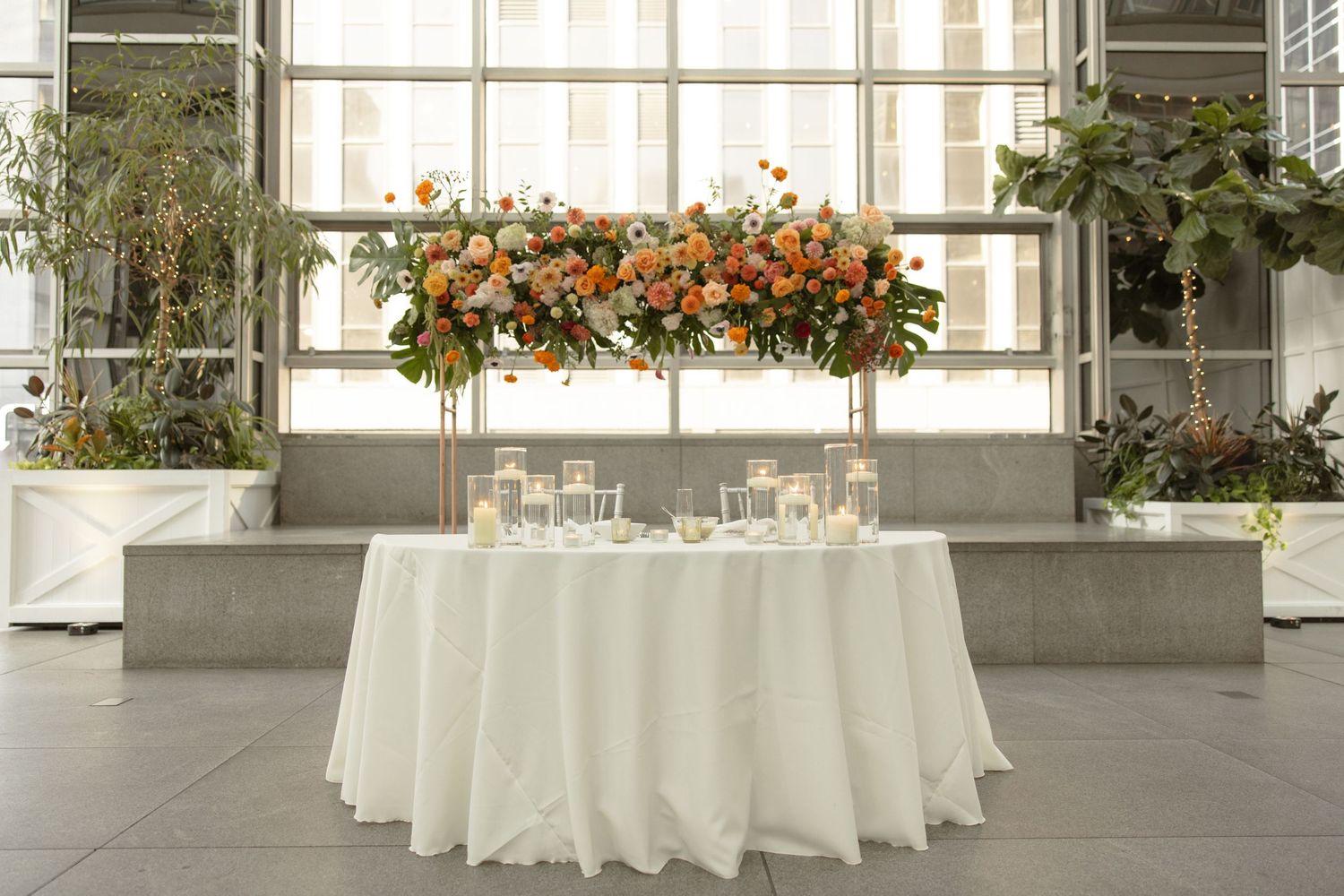 Reception table setup with white linens and hanging floral arrangement in orange and peach tones.