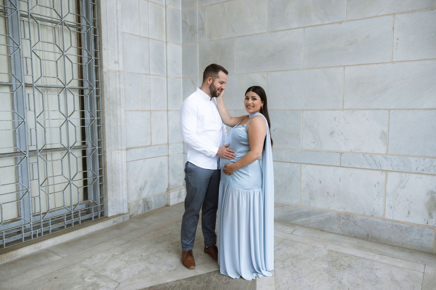 A couple in elegant attire share tender moments during a romantic photo session against a white stone wall and window.