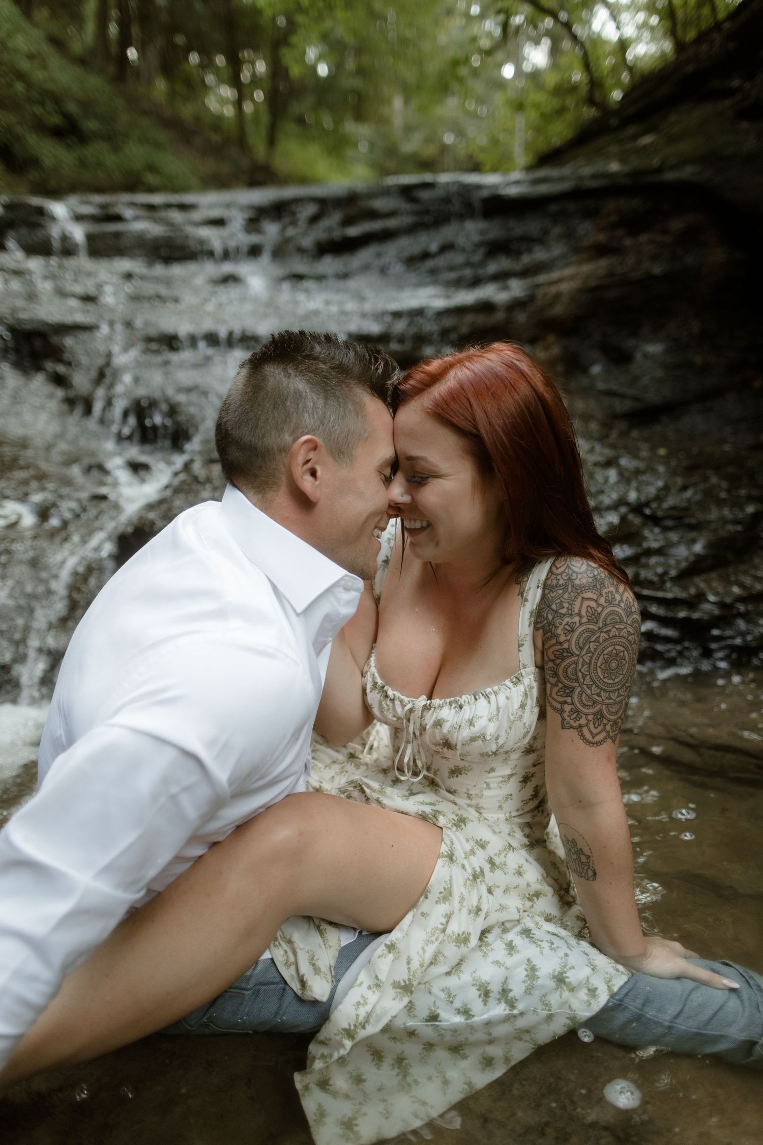 Romantic couple shares an intimate moment sitting by a cascading waterfall in a forest setting.