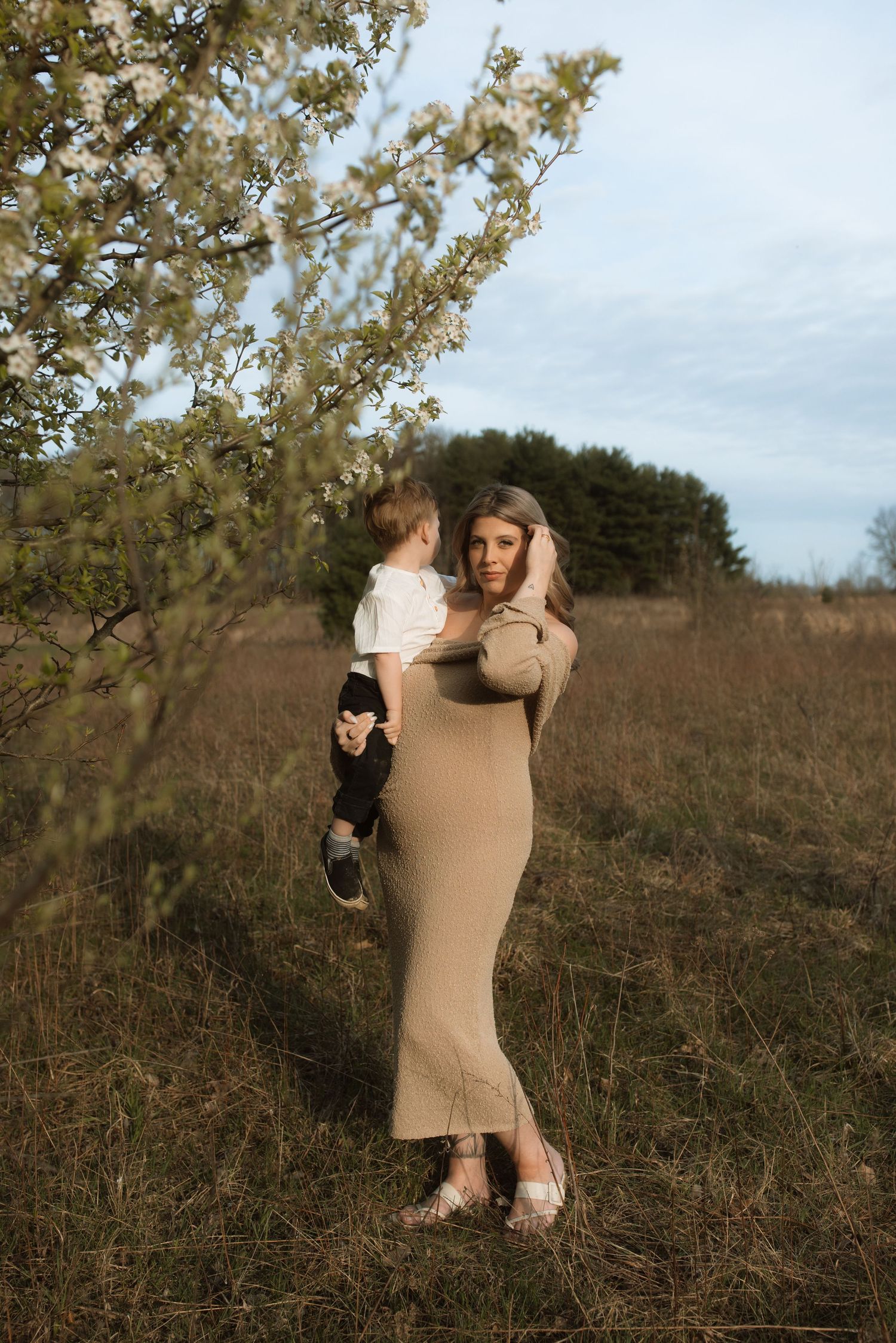 Mother and child standing together in field with blooming trees at sunset.