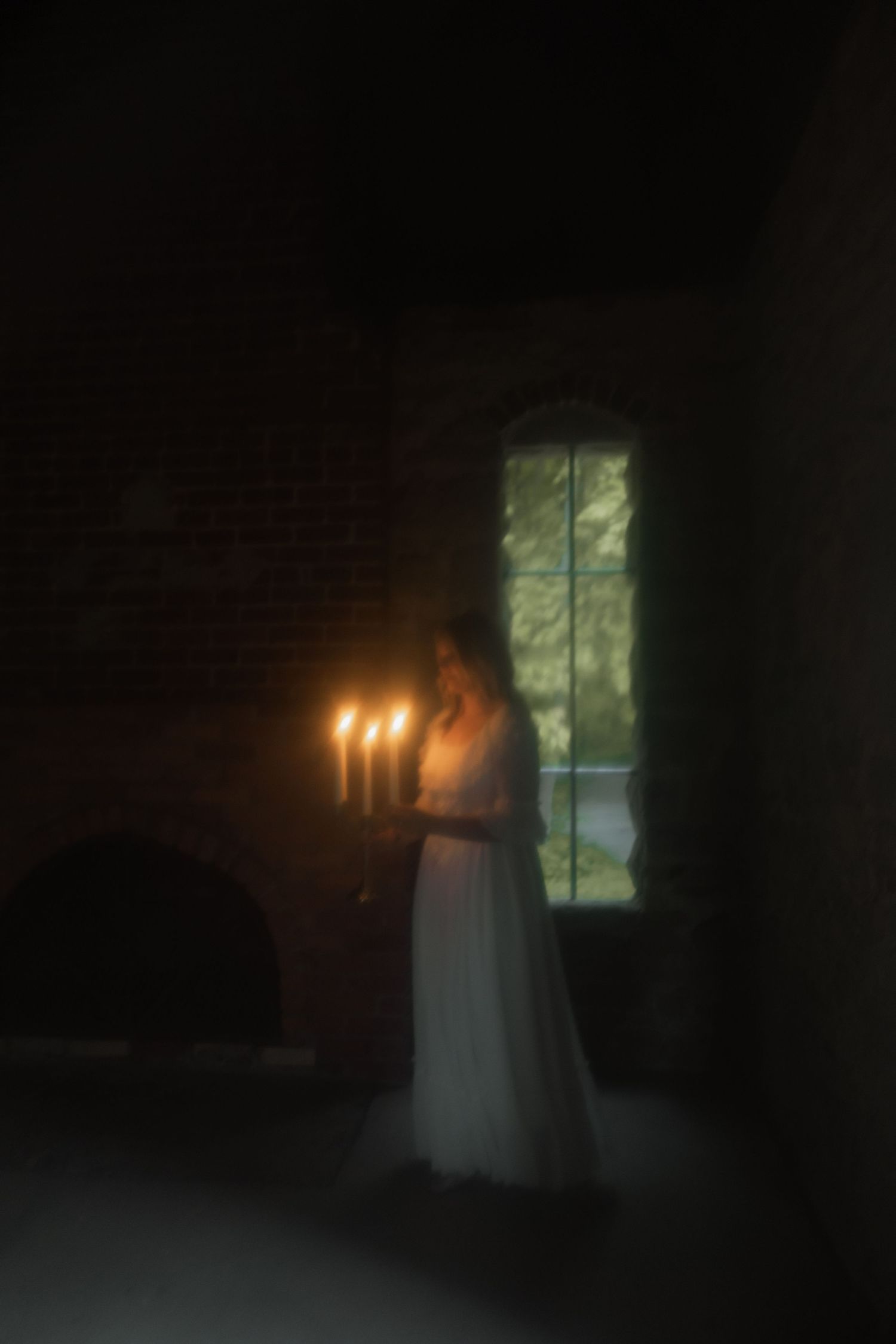 A figure in a white dress stands in a dark room holding lit candles near a window with a soft, ethereal glow.