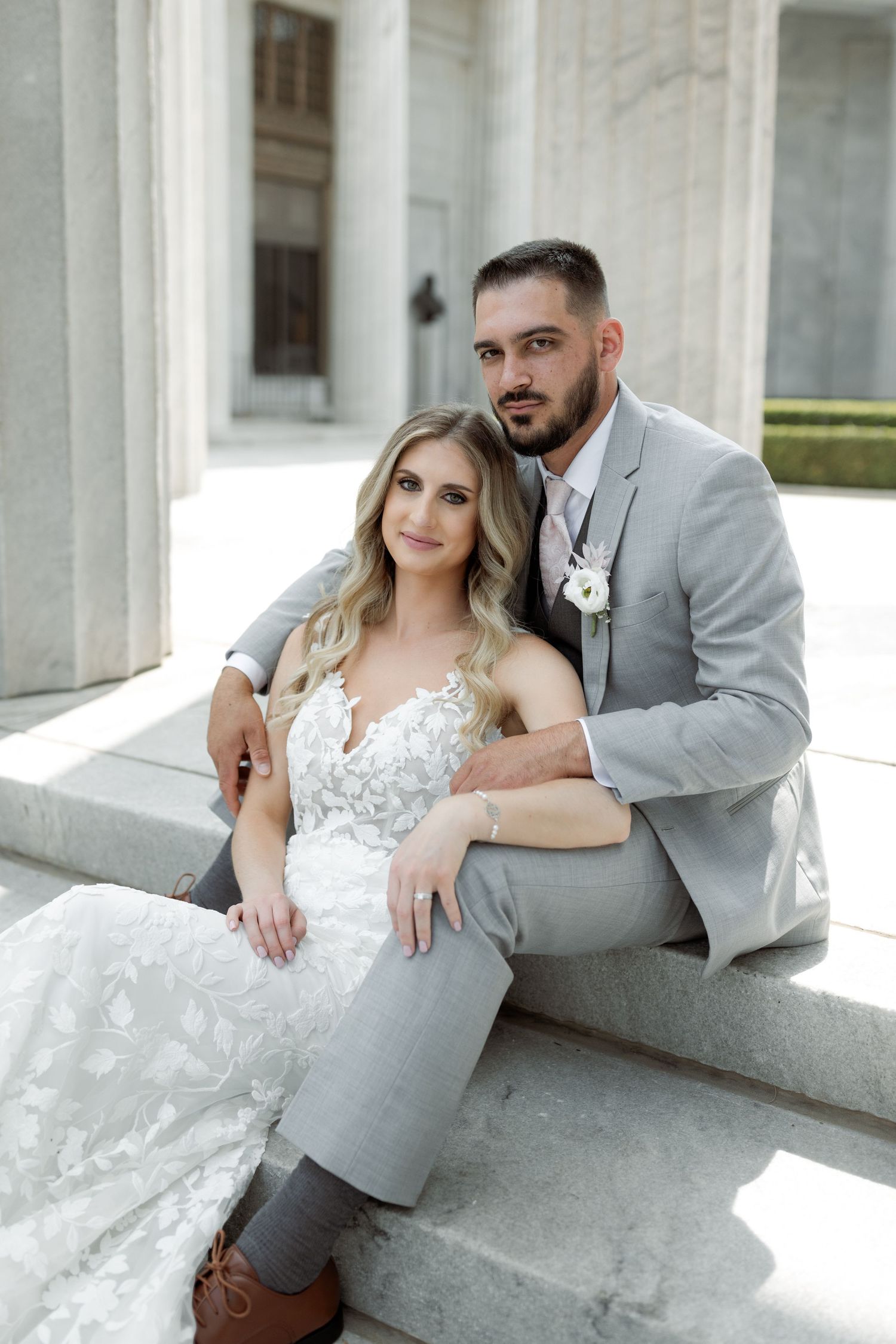 Newlyweds sitting on marble steps of classical building in grey suit and white lace gown.