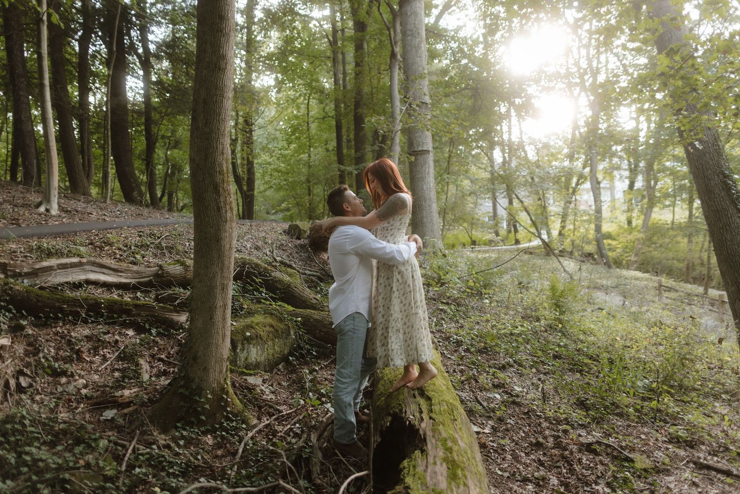 A romantic forest scene with sunlight streaming through trees while a couple embraces on a fallen log.