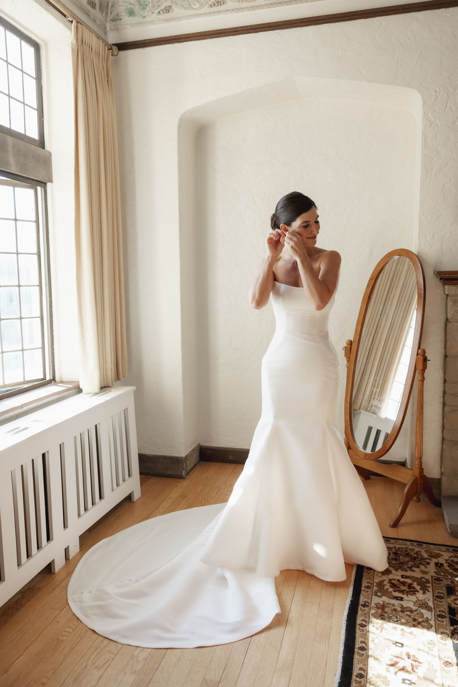 Elegant white mermaid wedding gown with sweeping train shown in bright sunlit room with vintage floor mirror.